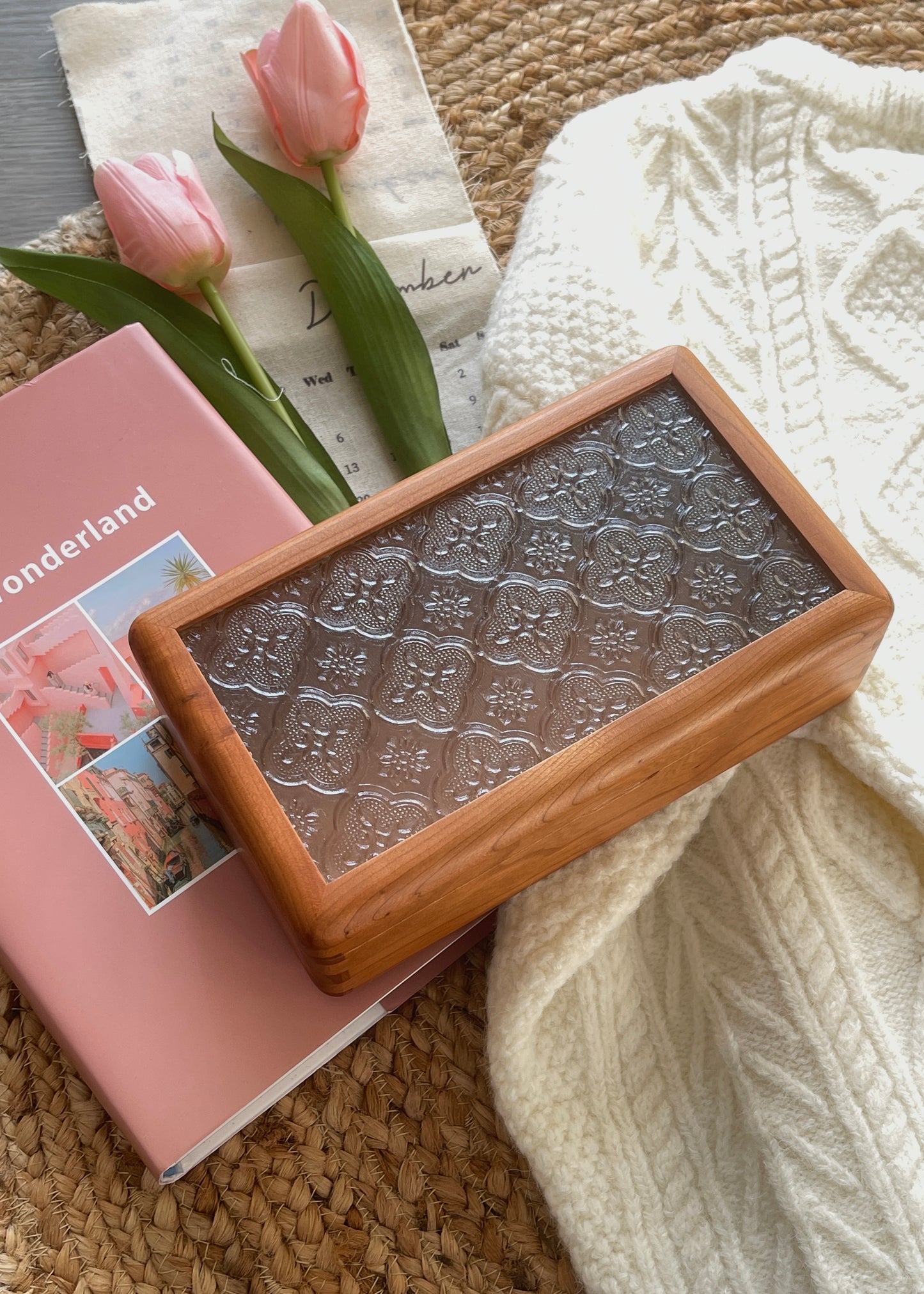 Wooden box with embossed design on a pink book with floral illustrations, next to pink tulips on a textured surface.